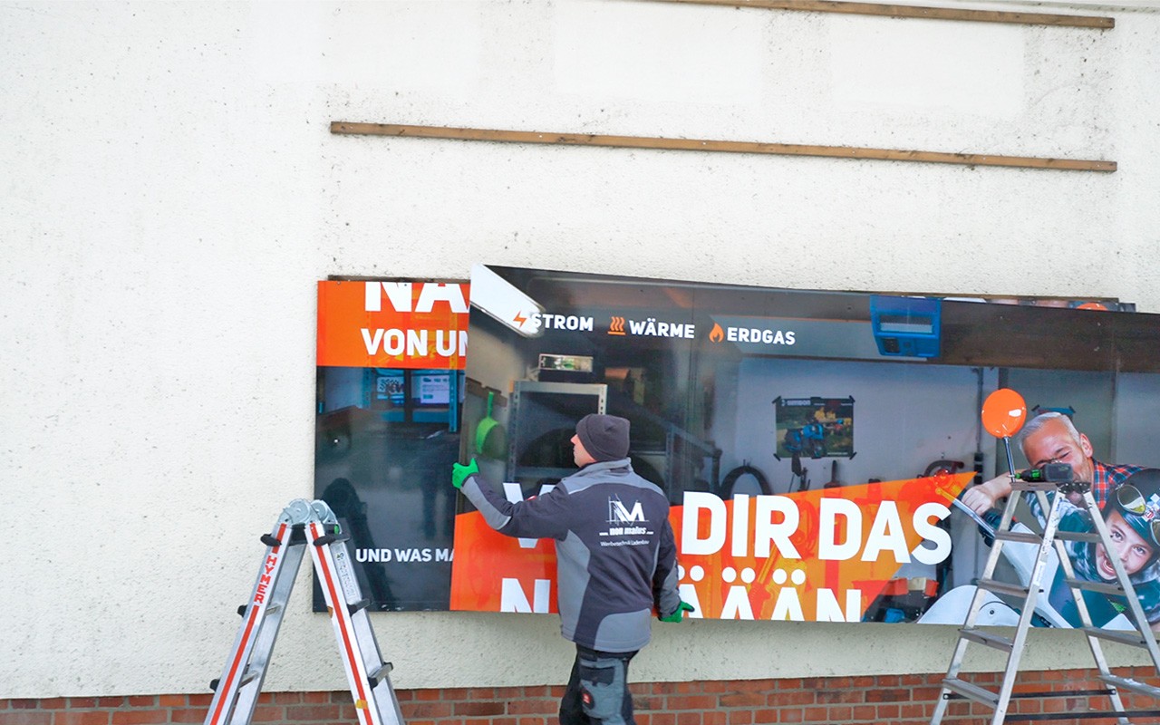Monteur trägt einen Teil eines großen Werbeschildes das gleich zu einem großen Werbeschild zusammengesetzt an der Wand angebracht wird